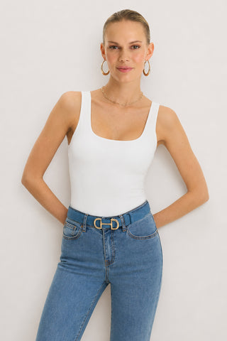 A woman stands against a plain background, smiling with her hands behind her back. She wears the Loren Bodysuit in white styled as a sleeveless top, paired with blue high-waisted jeans with a belt, gold hoop earrings, and a delicate necklace.