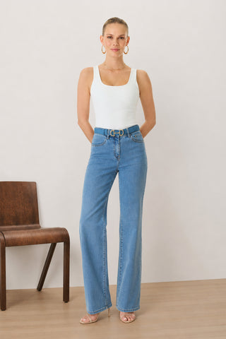 A woman stands indoors wearing the Loren Bodysuit in white—a versatile foundation piece—paired with blue high-waisted jeans featuring a belt and nude heels. Her hair is pulled back as she stands next to a brown wooden chair on a light floor.