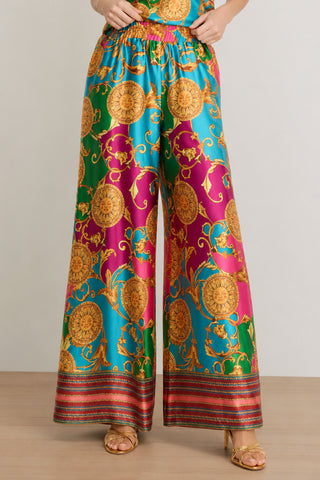 A person wearing the Marcia Silk Pant - Dolce Sun, featuring a vibrant baroque pattern in gold, teal, magenta, and green with a bold designer contrast print, paired with gold open-toe heels and standing on a wooden floor.