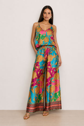 A woman stands elegantly against a plain light background, wearing a colorful sleeveless top paired with the Marcia Silk Pant - Dolce Sun, which features ornate gold patterns on blue, pink, and green silk fabric. The striking designer contrast print of the pants makes them perfect for summer.
