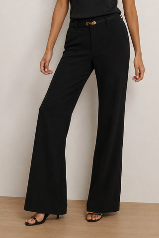 A woman models the Martina Belted Trouser in black, featuring a high-rise waist and flare leg silhouette, paired with black heeled sandals against a neutral background.