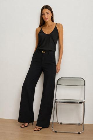 A woman with long straight hair stands beside a clear folding chair on a light wood floor, against a simple white background. She is wearing black heeled sandals and the Martina Belted Trouser - Black, which features a wide-leg silhouette and a high-rise waist, paired with a black camisole.