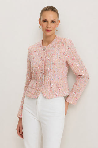 A woman stands against a plain light background wearing the May Tweed Crystal Jacket in Pink Multi, featuring small floral details, paired with white pants. Her hair is pulled back and she accessorizes with hoop earrings.
