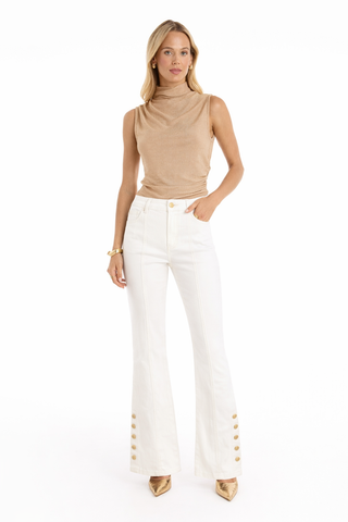 A woman stands against a white background wearing the Mia Shimmer Tank in champagne, high-waisted white flare pants with gold ankle buttons, gold pointed-toe shoes, and a gold bracelet.