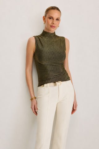 A woman styled in the Mia Shimmer Tank - Gold, a sleeveless, metallic turtleneck evening top, paired with high-waisted cream pants and accessorized with gold earrings, bracelets, and a belt, stands against a plain light background.