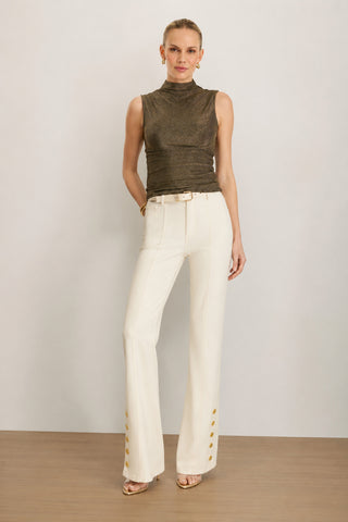 A woman stands against a neutral background, wearing the Mia Shimmer Tank in gold, paired with high-waisted white pants featuring gold buttons at the hem, a white belt, gold earrings, and heeled sandals.