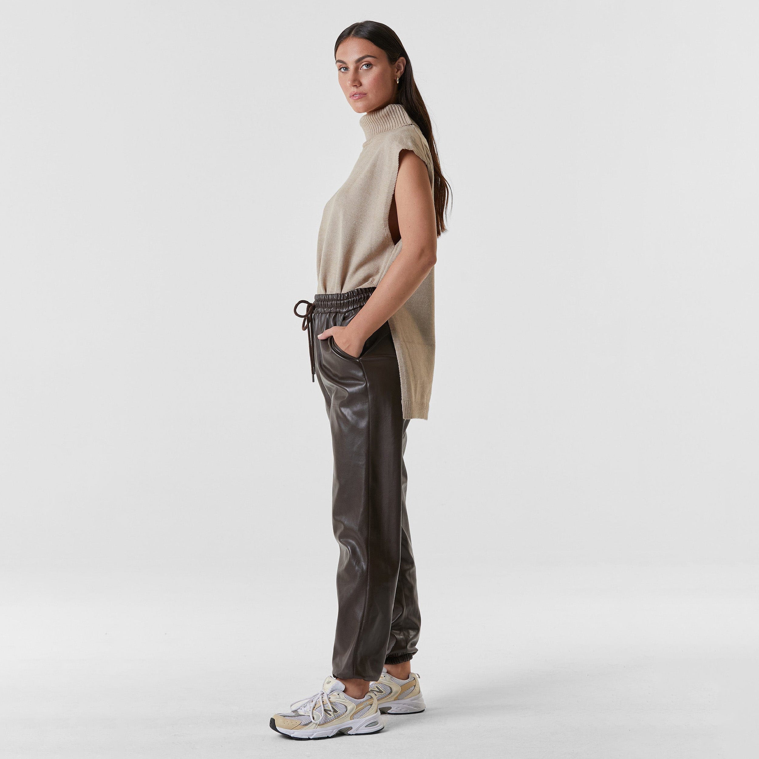 Side view of woman wearing dark brown high rise faux leather jogger with drawstring waistband and cinched ankles with suede lining.