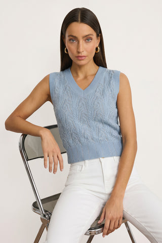 A woman with long brown hair sits on a metal folding chair, wearing the Mikaela Crystal Knit Top in sky blue and high-waisted white pants, looking directly at the camera against a plain background.