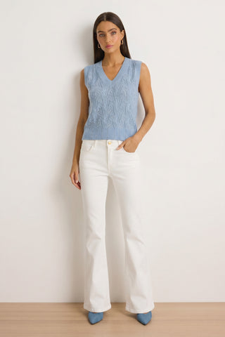A woman with straight brown hair and a neutral expression stands against a white wall wearing the Mikaela Crystal Knit Top in sky blue, styled with white high-waisted flared pants and blue pointed-toe heels.