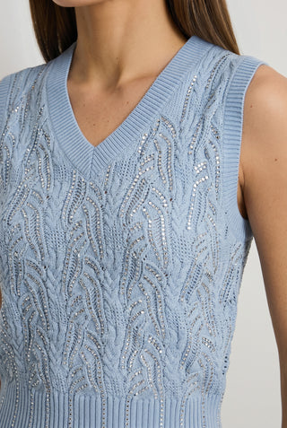 A woman wears the Mikaela Crystal Knit Top in sky blue, featuring a sleeveless V-neck design with intricate textured patterns and shiny silver accents.