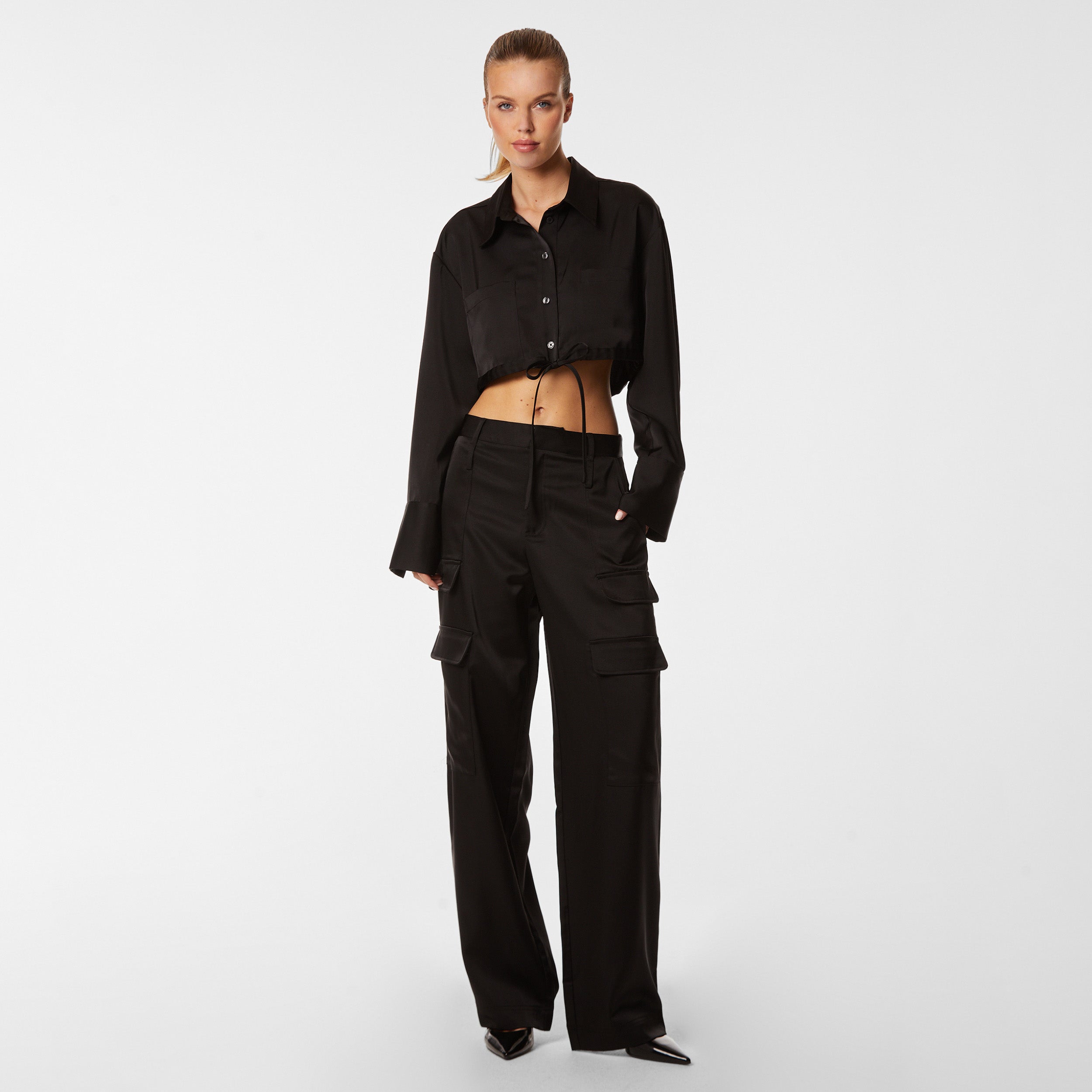 Full body view of woman wearing black Milan Satin Button Up features luxurious and structured satin-like fabric, featuring cargo detailing, shell buttons and a tie-waist