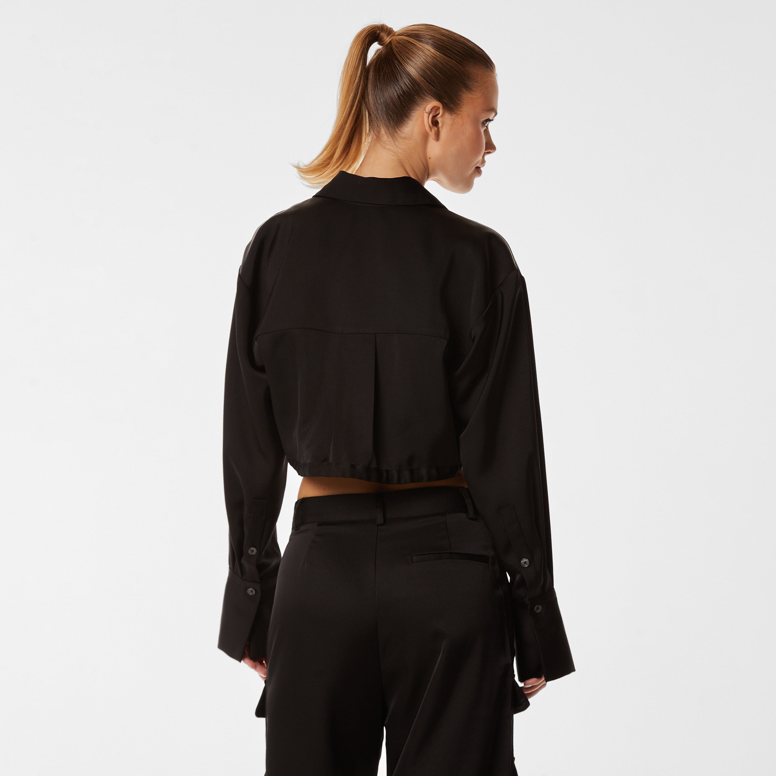 Rear view of woman wearing black Milan Satin Button Up features luxurious and structured satin-like fabric, featuring cargo detailing, shell buttons and a tie-waist
