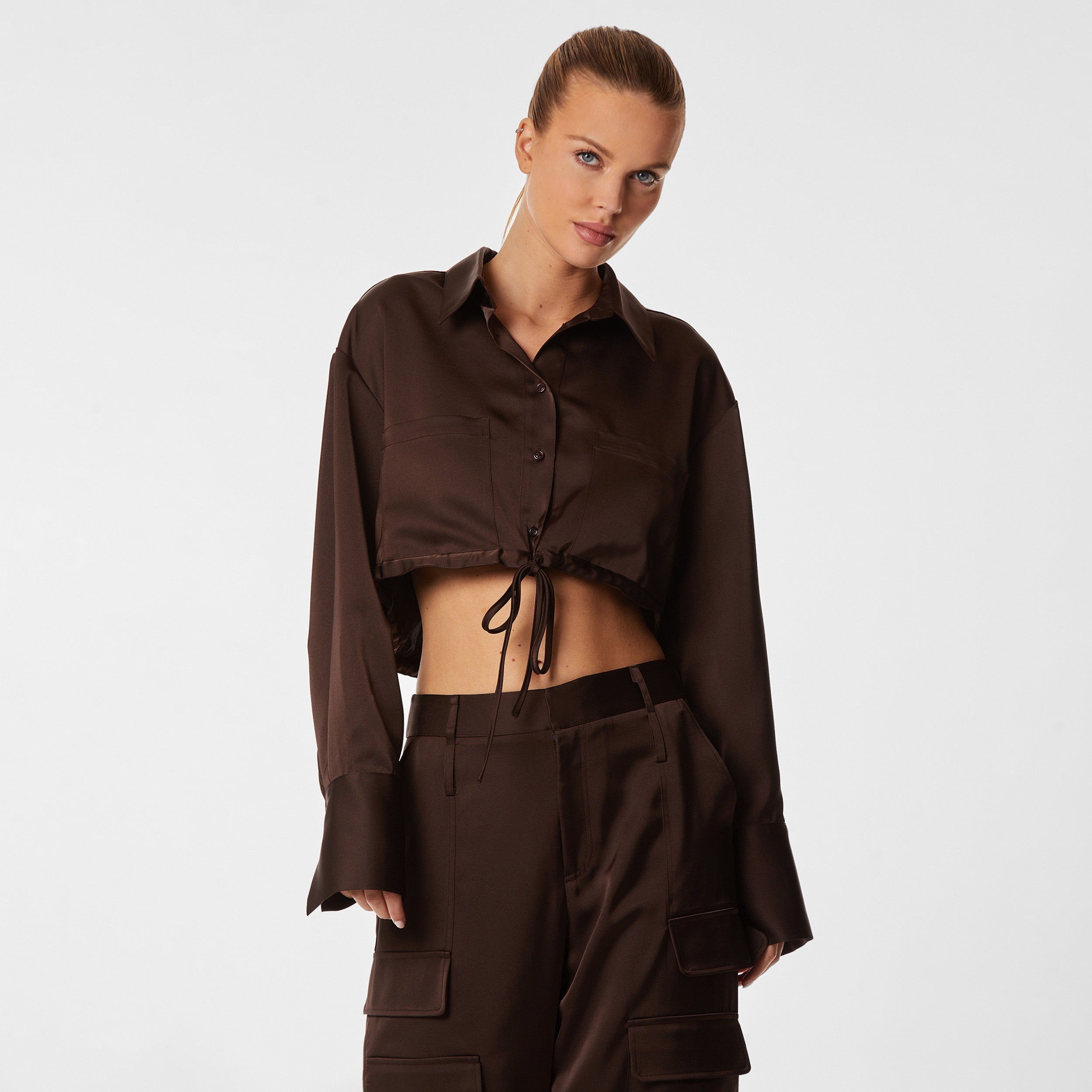 Front view of woman wearing brown Milan Satin Button Up features luxurious and structured satin-like fabric, featuring cargo detailing, shell buttons and a tie-waist