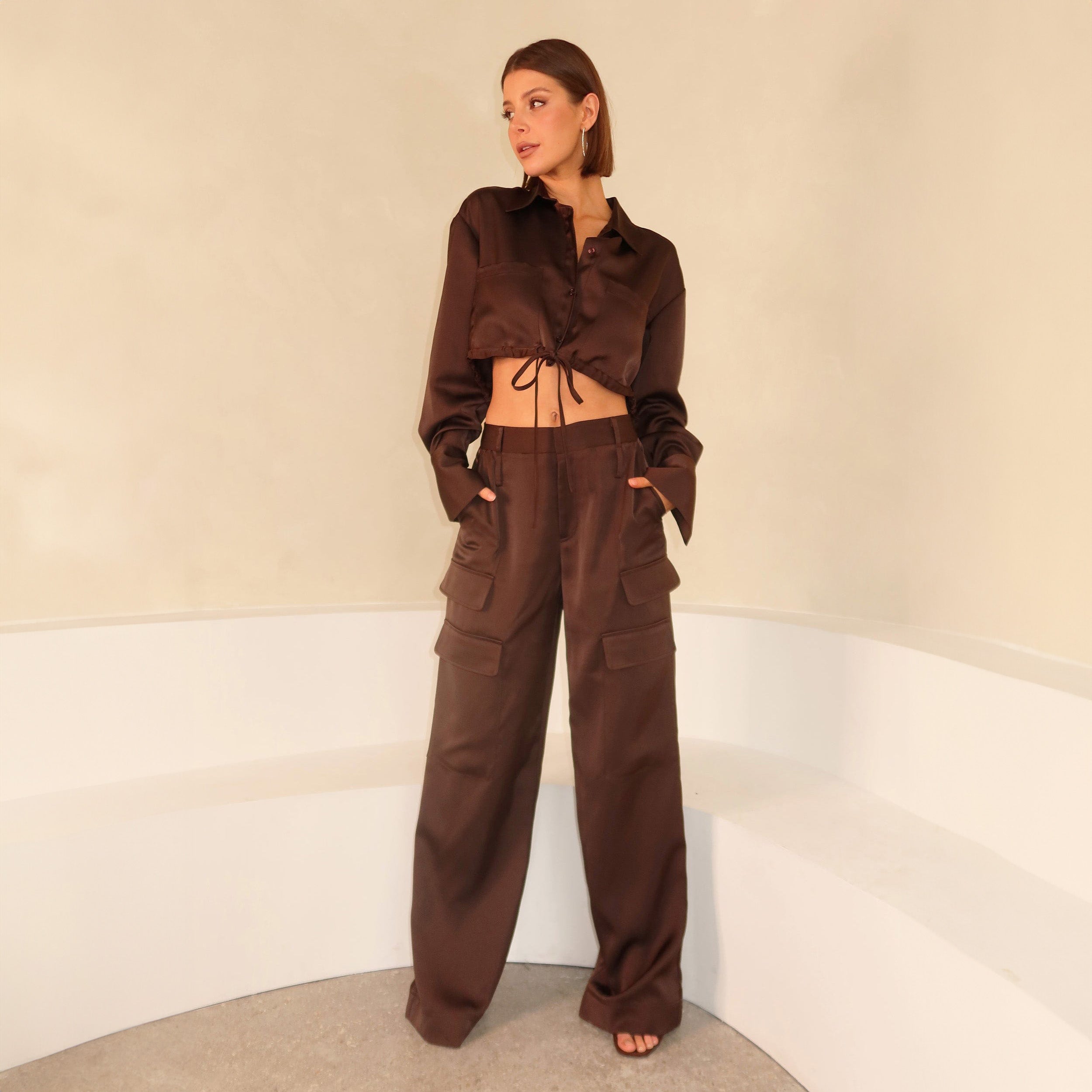Full view of woman wearing Brown Satin Milan Pant features Mid-rise with side pockets and cargo detail. Luxurious and structured satin-like fabric. 