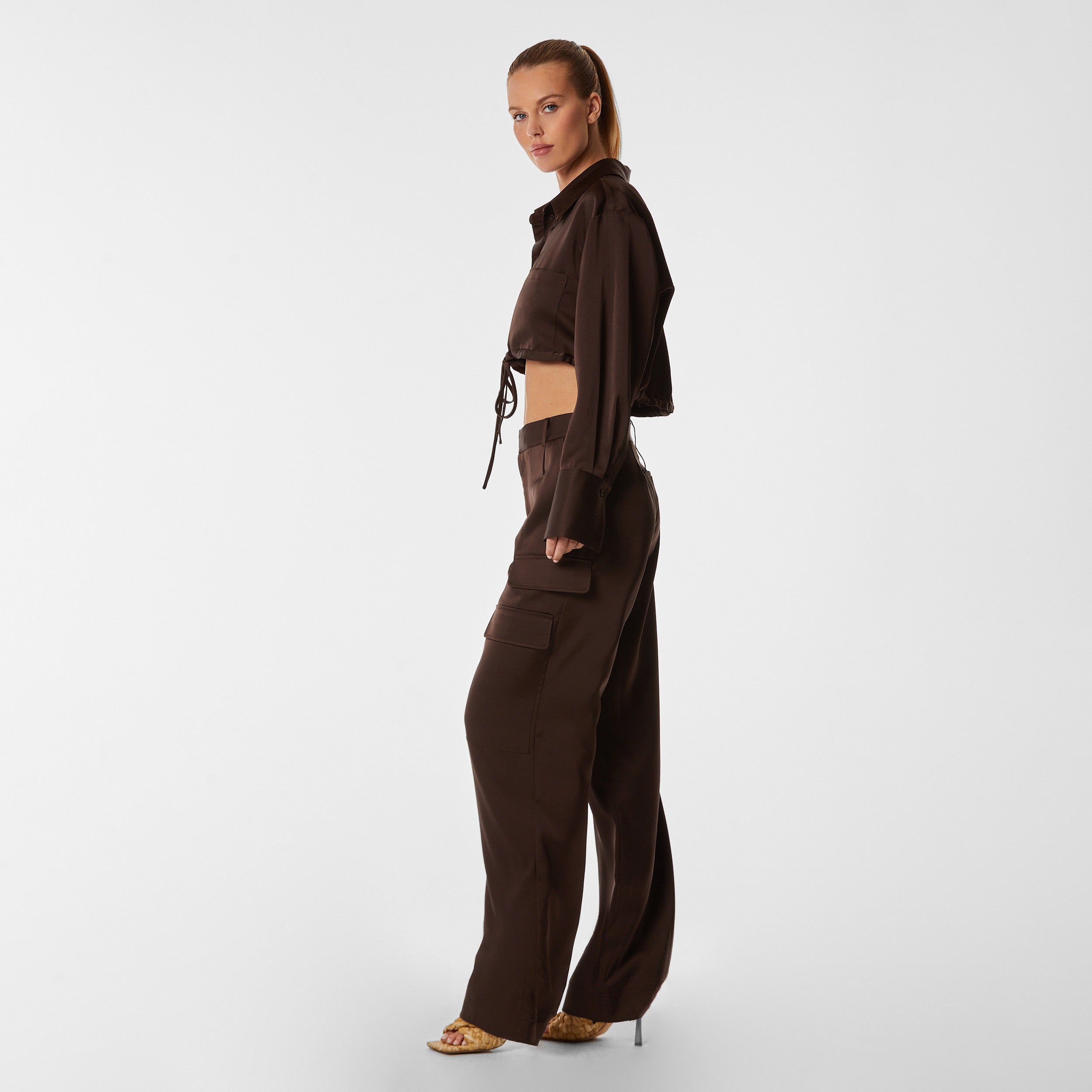 Side view of woman wearing Brown Satin Milan Pant features Mid-rise with side pockets and cargo detail. Luxurious and structured satin-like fabric. 