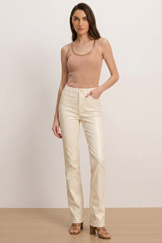 A woman stands against a plain background, wearing the Millie Knit Tank - Mocha—a ribbed, sleeveless mocha top with chain detail—paired with cream straight-leg pants and tan heeled sandals. She gazes forward with one hand in her pocket.