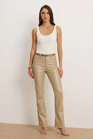A woman stands against a plain white wall, wearing the Millie Knit Tank - Pearl with a gold chain neckline, beige metallic pants, clear-heeled sandals, gold hoop earrings, and a bracelet.