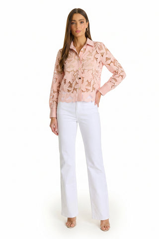 A woman models the Mina Lace Blouse in blush, featuring long sleeves and delicate floral lace, paired with high-waisted white pants and nude heels against a plain white background.