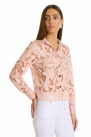 A woman models the Mina Lace Blouse - Blush, a light pink long-sleeve lace top with floral patterns and buttons, paired with white pants, standing against a plain white background and looking slightly to the side.