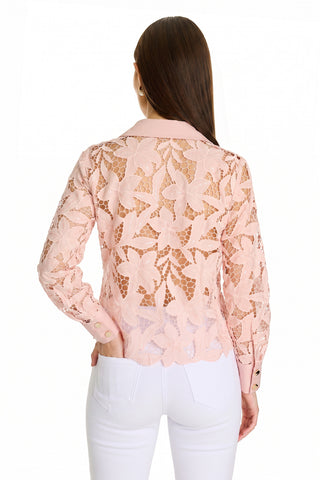 A woman with long brown hair stands facing away, wearing the Mina Lace Blouse - Blush featuring delicate floral lace and white pants, with one hand in her pocket.