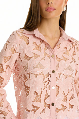 A woman wears the Mina Lace Blouse - Blush, a light pink, semi-sheer lace top with floral patterns and shiny buttons. Paired with pearl earrings and natural makeup, the collared blouse adds a delicate touch to her look.