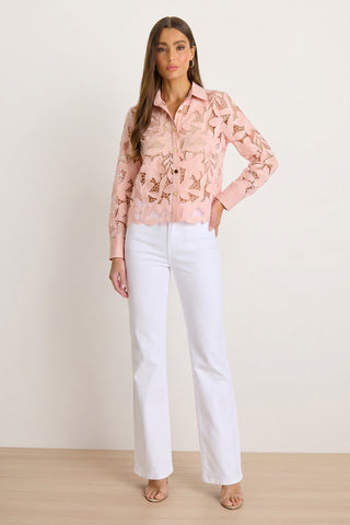 A woman stands indoors wearing the Mina Lace Blouse in blush, featuring long sleeves and floral lace, paired with white high-waisted wide-leg pants. She has long brown hair and nude heels.