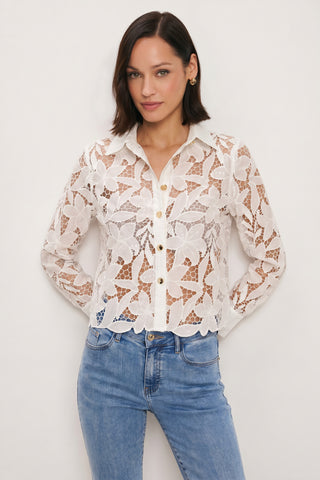 A woman wearing the Mina Lace Blouse in white with a delicate floral pattern and light blue jeans stands against a plain white background, hands behind her back.