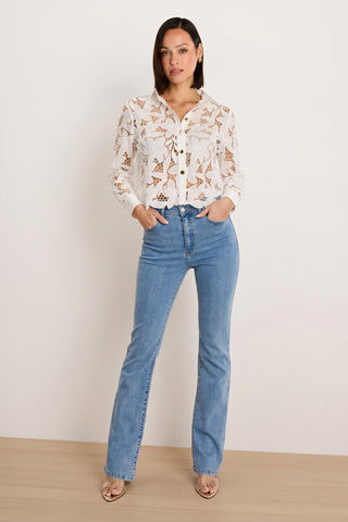 A woman stands against a plain background wearing the Mina Lace Blouse - White over a nude cami, paired with light blue high-waisted jeans and beige heeled sandals, hands in her pockets.