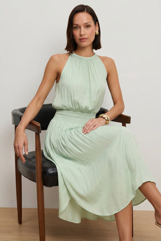 A woman with shoulder-length brown hair wears the Emelia Halter Dress - Mint while seated on a black and wood chair against a plain background, styled with gold earrings and a bracelet.