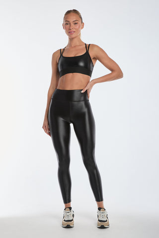 Wearing a Liquid Jolie Bra in Black Gloss and matching leggings, paired with sleek white and black sneakers, the person strikes a confident pose against a plain white backdrop, one hand on their hip.