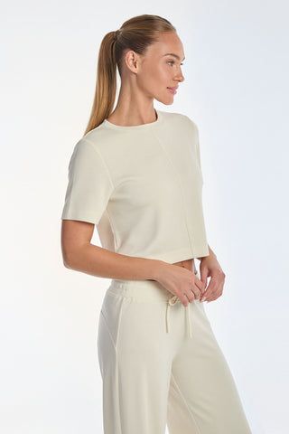 A person with long hair in a ponytail is wearing the Daphne Soft T-Shirt - Pearl, paired with matching pants. The ultra-soft fabric complements their relaxed posture as they stand against a plain light background, gazing to the side.