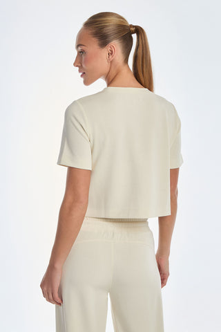 A person with long hair in a ponytail wears the Daphne Soft T-Shirt - Pearl, paired with matching pants on an ultra-soft fabric, in light beige. They face slightly away from the camera, gazing to the side against a plain white background.