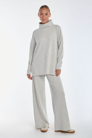 A person confidently poses against a plain white background, wearing a cozy light grey oversized turtleneck sweater and Heather Grey Daphne Pant in a soft modal blend, epitomizing luxurious comfort.