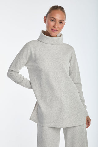 A person in a Daphne Top - Heather Grey turtleneck and matching pants stands against a plain white background. The luxurious comfort and subtle sheen of the soft modal blend complement their gentle smile, with one arm behind their back and neatly tied hair.
