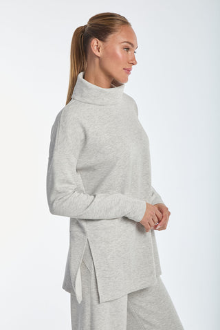 A woman in the Daphne Top - Heather Grey turtleneck sweater and matching pants of soft modal blend stands against a plain background. Her hair is in a ponytail, and she looks to the side with a slight smile. The sweater features side slits and offers luxurious comfort with its relaxed fit.
