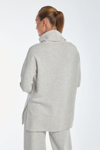 Standing against a plain background, a person exudes luxurious comfort in a light gray Daphne Top - Heather Grey turtleneck sweater. Crafted from a silky, breathable soft modal blend and paired with matching pants, the ensemble beautifully complements their tied-back hair as they face away.