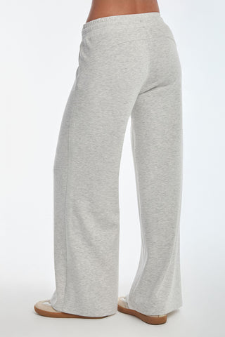 Wearing the Daphne Pant in Heather Grey, made from a soft modal blend for luxurious comfort, a person stands gracefully on a white surface. The wide-leg fit falls near their shoes, seen from behind.