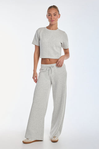 A person stands against a white background wearing the Daphne Soft T-Shirt in Heather Grey, paired with matching wide-leg pants. Their hair is tied back, and they're sporting white sneakers.