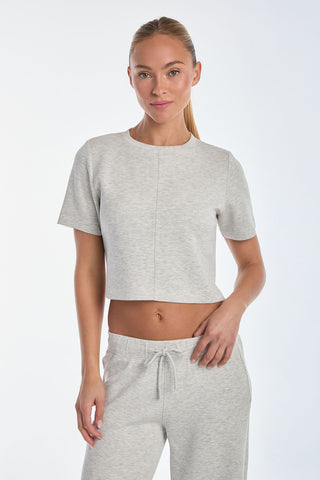 Someone with long hair is donning the Daphne Soft T-Shirt in Heather Grey and complementary joggers, made from ultra-soft fabric. They pose against a plain white background, facing the camera with one hand on their hip.