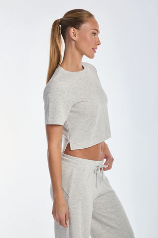 A person with long hair tied back is wearing the ultra-soft Daphne Soft T-Shirt in Heather Grey, paired with matching sweatpants, standing in a side profile against a plain white background.