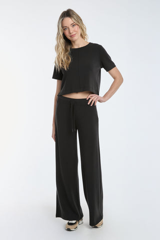 A person with long, wavy hair models the black Daphne Soft T-Shirt, featuring a short-sleeve, crop top style. Paired with wide-leg pants and striking a confident pose against a plain white backdrop, they express a laid-back vibe.
