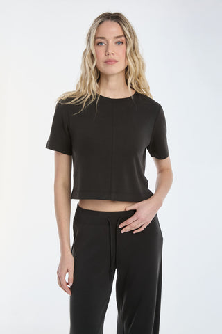 A long-haired person in a fitted Daphne Soft T-Shirt - Black and black pants stands against a plain background, looking at the camera.