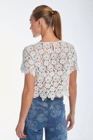 A woman with a ponytail, slightly turned from the camera, is wearing a white Sadie Lace T-Shirt with floral lace and blue jeans. The light, neutral background perfectly complements her spring-ready look.
