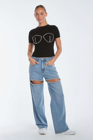 A person wearing the Rya Crystal T-Shirt - Black with hand-beaded crystals on the front, paired with wide-leg blue jeans featuring open thigh sections. Their hands are in their pockets, standing against a plain white background.