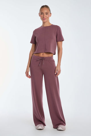 A person wears the Daphne Soft T-Shirt in Orchid, a mauve two-piece lounge set with a short-sleeve cropped top and wide-leg pants, against a plain white background. They finish the look with crisp white sneakers.