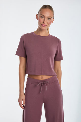 A person is standing against a plain background, wearing the Daphne Soft T-Shirt in Orchid and matching pants. The outfit boasts an ultra-soft fabric, minimalist design, short sleeves, and drawstring pants. The person has a neutral expression.
