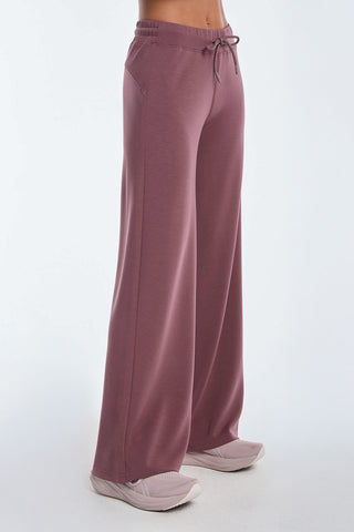 A person wears Daphne Pant - Orchid, wide-leg and in a luxurious comfort modal blend with a drawstring waistband, paired with light pink sneakers. The plain white background focuses on the side view of the lower body—a look perfect for travel.