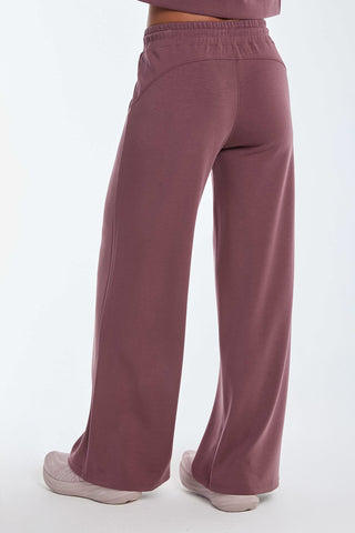 A person models the Daphne Pant - Orchid, in wide-leg style and crafted from a soft modal blend, paired with white sneakers against a neutral backdrop. The rear view highlights the relaxed fit and elastic waistband, providing luxurious comfort ideal for travel.