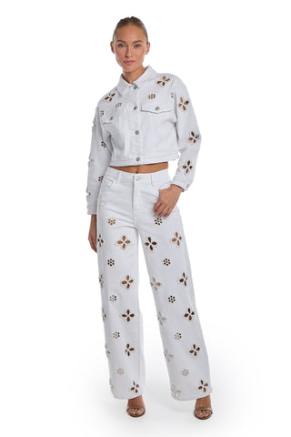 A woman poses with one hand in her pocket, wearing the Kaia Pearl High Rise Jean in white and a matching stretch denim jacket, both featuring cut-out floral designs. Her hair is pulled back, and she completes the look with open-toed heels.