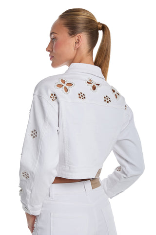 A woman wears the Kaia Pearl Jean Jacket in white, featuring floral cut-out details on the back and sleeves, paired with high-waisted white pants. She stands facing away, head turned to the side, her hair in a sleek ponytail.